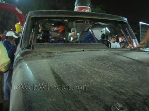 Dusty hood and windshield of a race truck with driver in helmet visible at night
