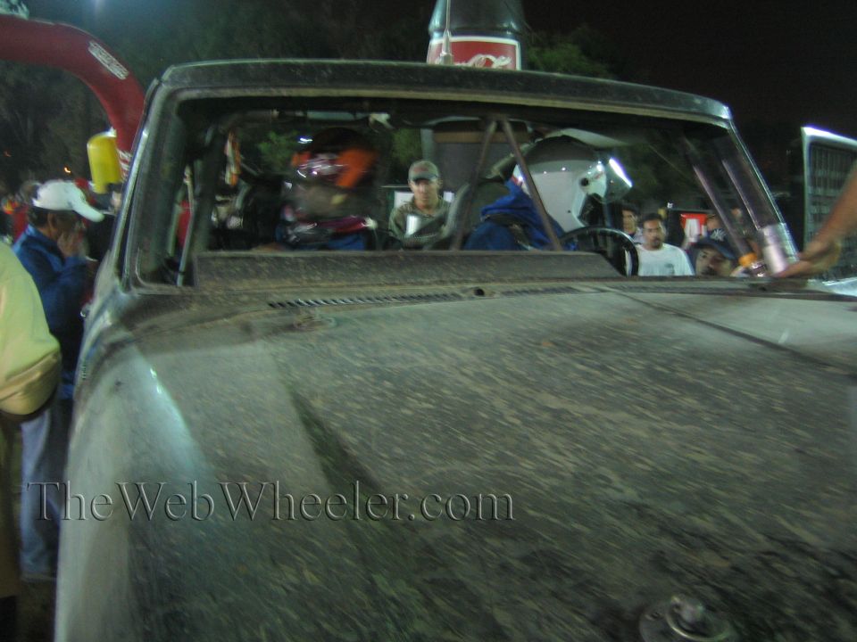 Dusty hood and windshield of a race truck with driver in helmet visible at night