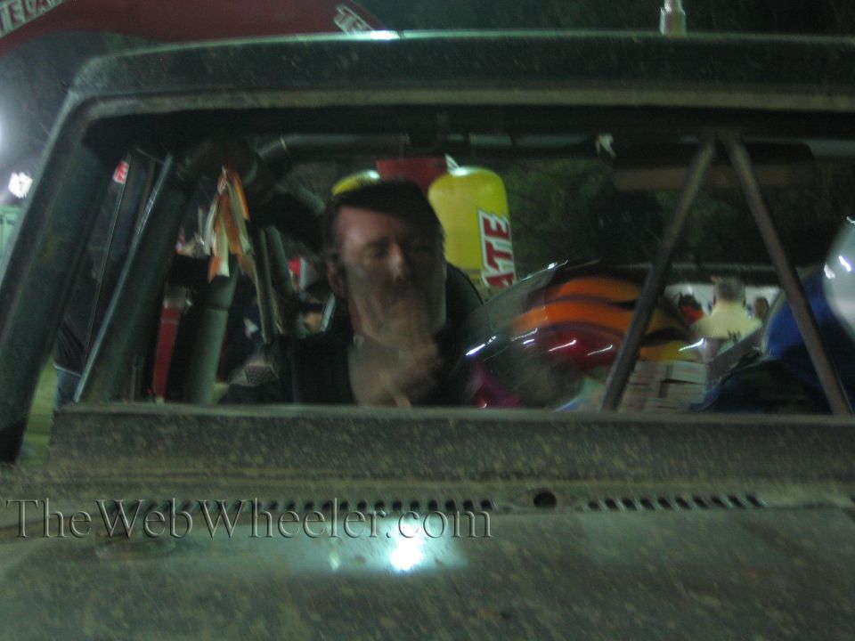 Driver gesturing inside the dusty cab of a race truck through the windshield at night