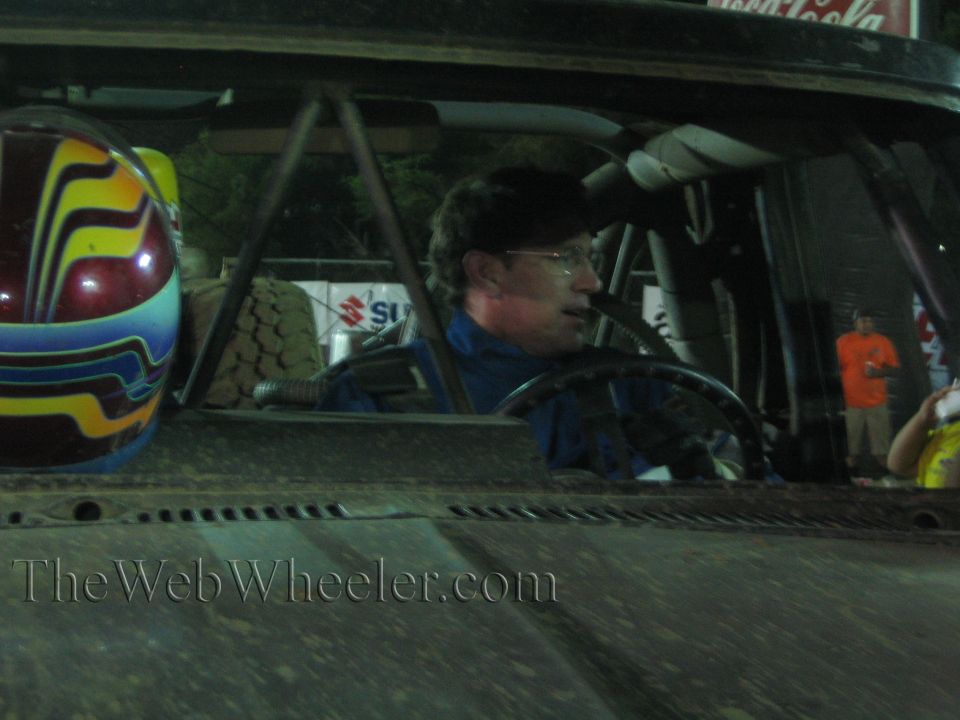 Bespectacled driver in blue suit at the wheel of a race truck with painted helmet at night