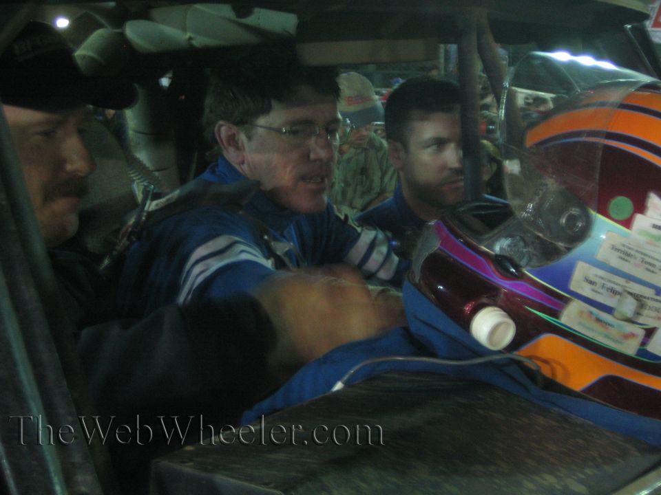 Men in blue race suits beside a colorful helmet with stickers inside a race truck at night