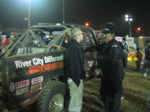 Two men talk beside black race truck with River City Differentials and West Coast decals at night