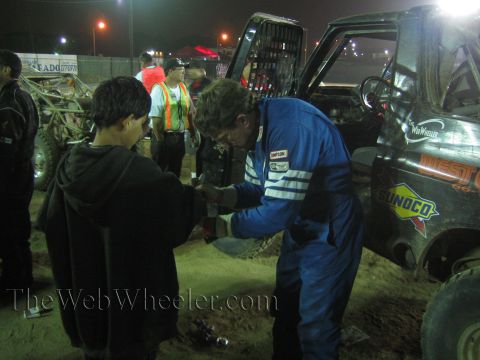 Man in blue racing suit shakes hands with a youth at a nighttime off-road pit