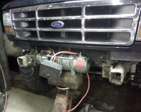 1995 Ford Bronco - Low front view of a dark blue Ford truck's chrome eggcrate grille with blue Ford oval, photographed in a dim garage with the front bumper r…
