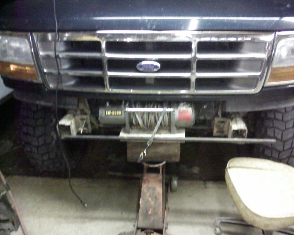 Dark blue Ford Bronco front bumper with chrome grille, headlights, and a winch mounted below. Tires visible