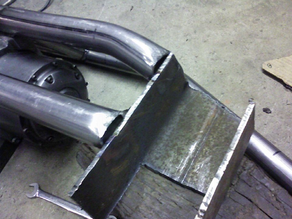 Custom exhaust pipe section welded to a metal bracket, with a differential visible behind