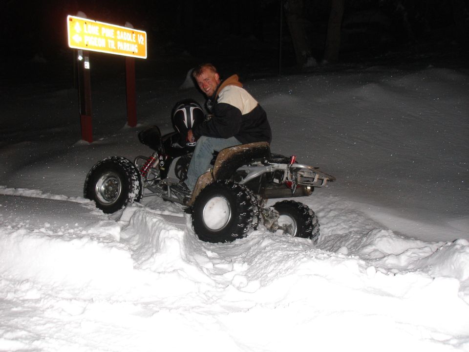 Rider seated on a sport quad ATV in deep snow at night beside a Lone Pine Saddle trail sign