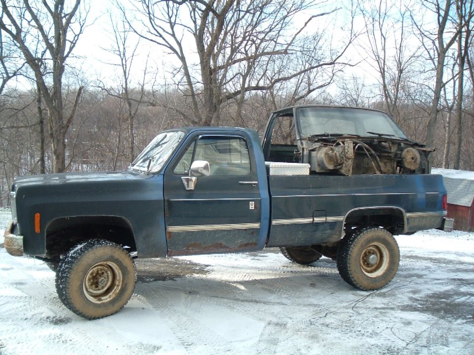 Dark blue pickup truck with a disassembled truck cab and engine parts in the bed, parked in snow. The truck appears to be from the 1970s