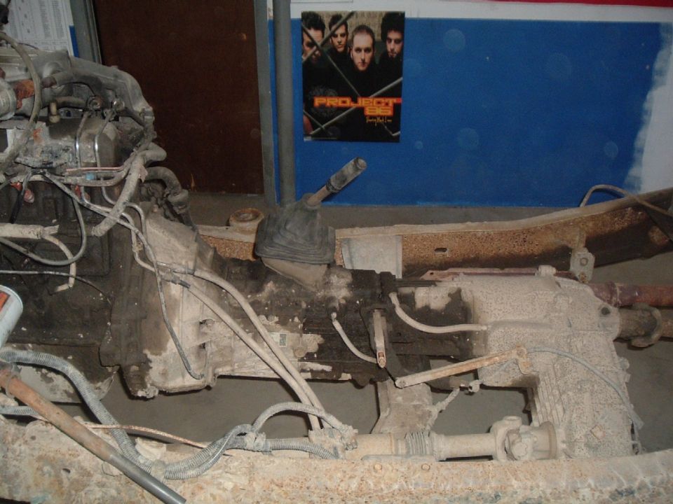 Truck chassis showing engine, transmission, and drivetrain components. A poster is visible on the wall in the background