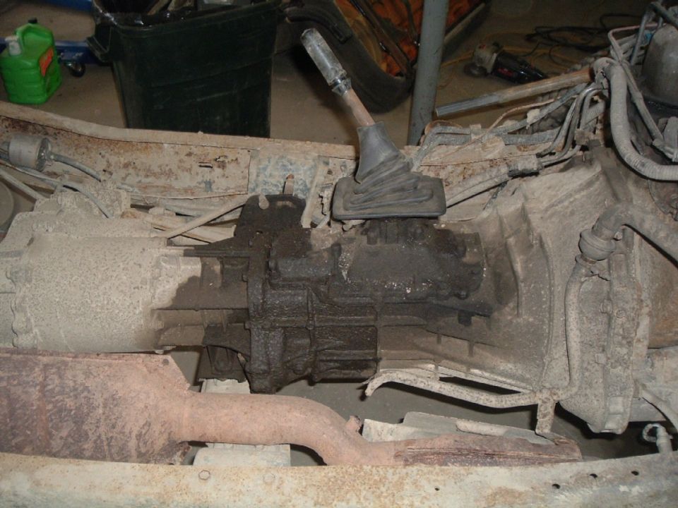 Transfer case and transmission linkage on rusty frame rails, with exhaust pipe visible