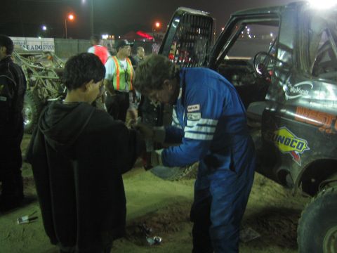 Driver in blue racing suit grips a youth's hand beside a Sunoco-decaled race truck at night