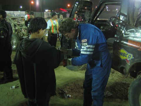 Bespectacled racer in blue Simpson suit greets a hooded youth at a night pit area
