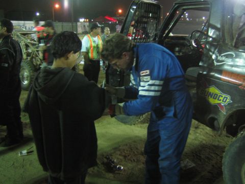 Curly-haired racer in blue suit clasps hands with a youth at a dirt race pit at night