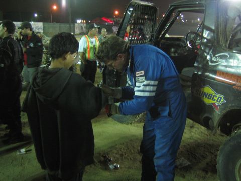 Bespectacled racer in blue suit greets a hooded youth in a dirt pit at night