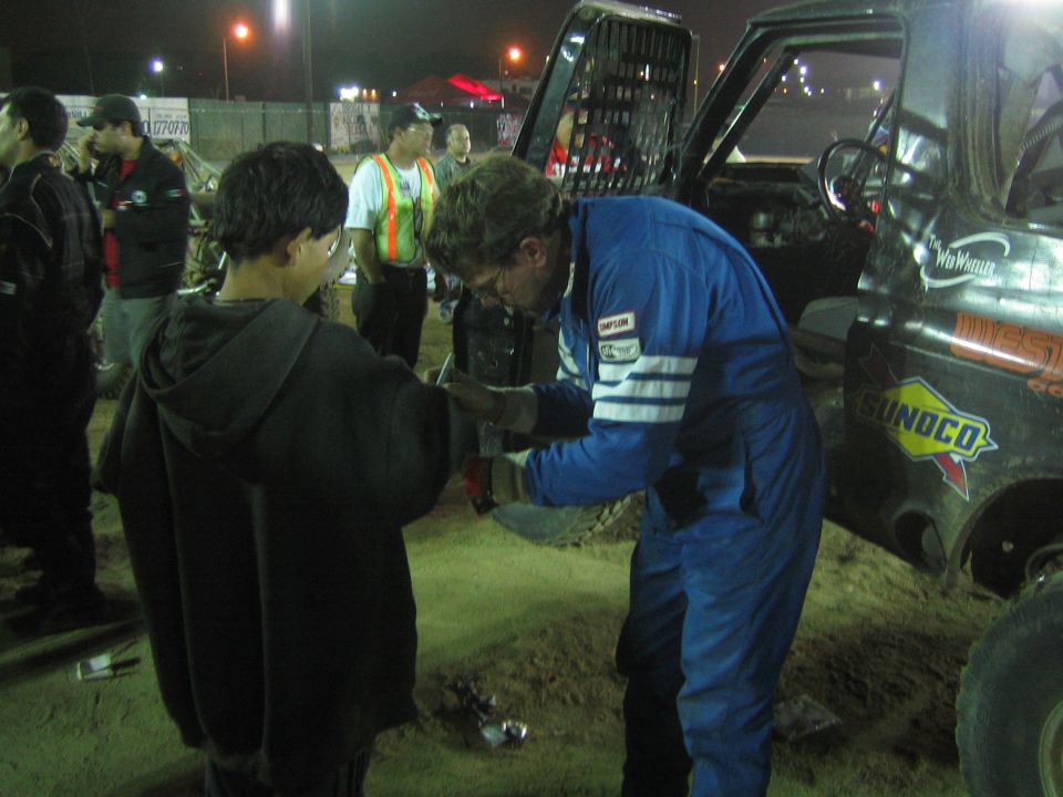 Bespectacled racer in blue suit greets a hooded youth in a dirt pit at night