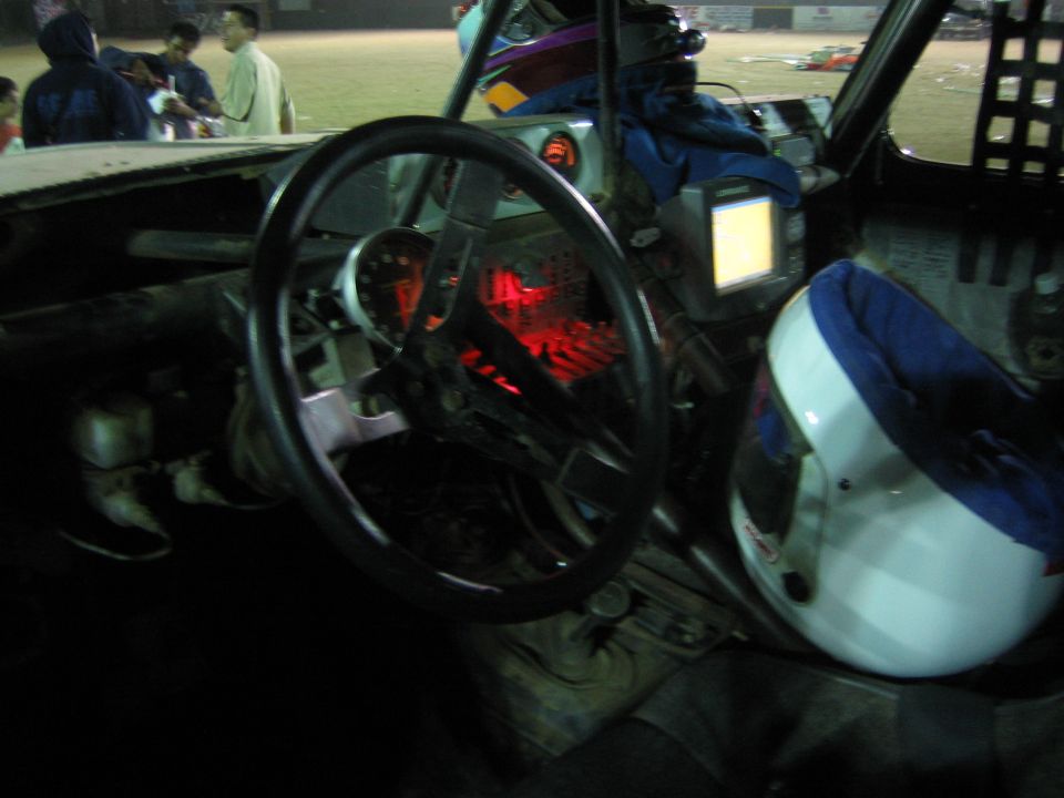 Race-truck interior with steering wheel and red-lit dash, people outside at night