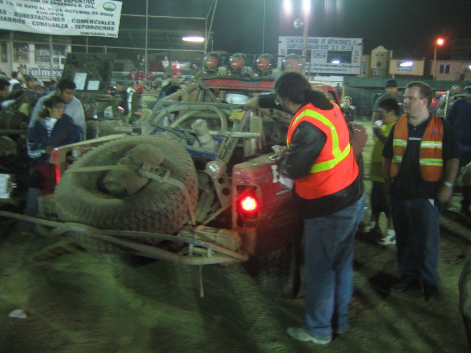 Official in orange vest works at the rear of a spare-tire-laden race truck at night