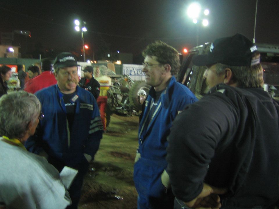 Men in blue racing suits and caps talk in a dirt pit area at night