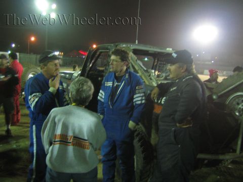Group in blue racing suits talk beside a race truck under night lights