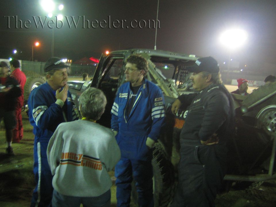 Group in blue racing suits talk beside a race truck under night lights