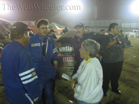 Racers in blue suits chat around a tire stack with sponsor decals at night