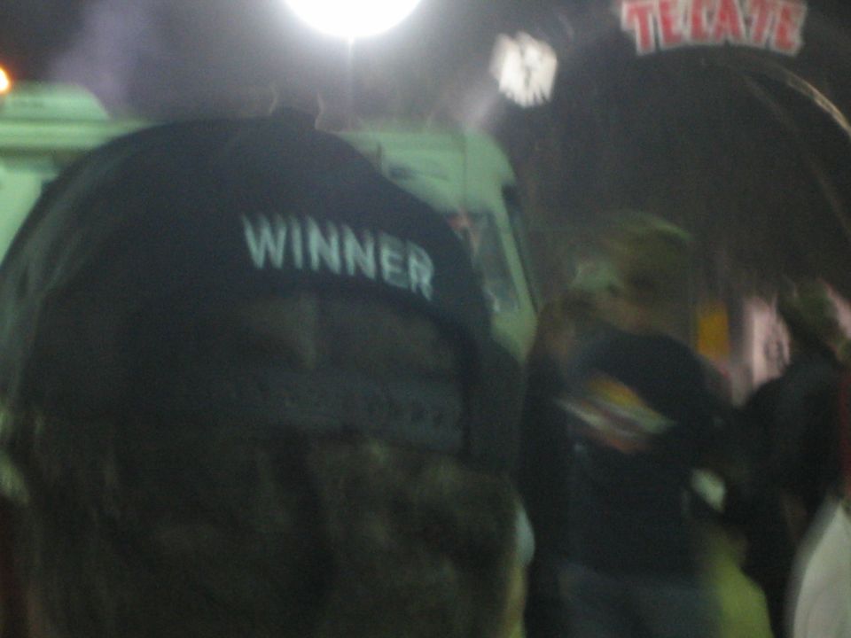 Back of a camouflage cap reading WINNER in a crowd under a Tecate sign at night