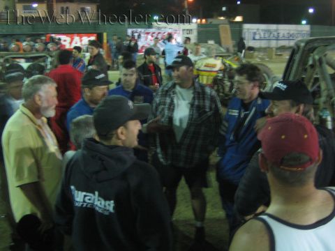 Crowd of men in caps and racing gear talk in a dirt pit under banners at night
