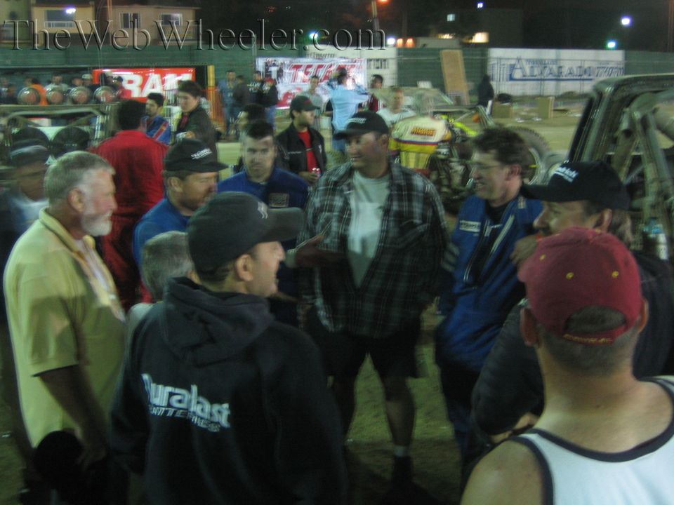 Crowd of men in caps and racing gear talk in a dirt pit under banners at night