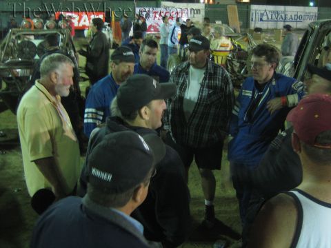 Group of men in caps and racing suits converse in a lit dirt pit at night