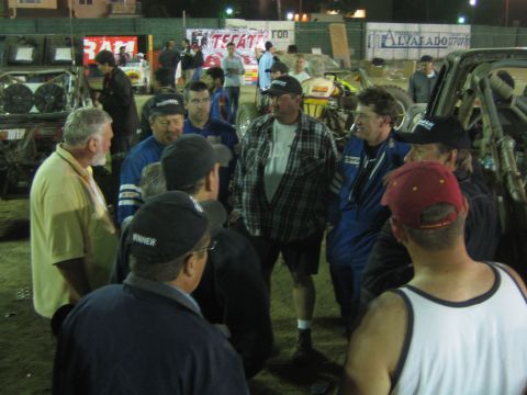 Group of men in racing suits and caps talking at night in a dirt pit area