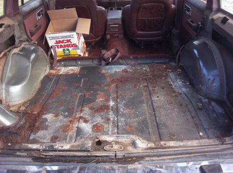 1987 Jeep Cherokee - The interior cargo area of a 1987 Jeep Cherokee shows significant rust on the floor, maroon upholstery, and a box of jack stands.