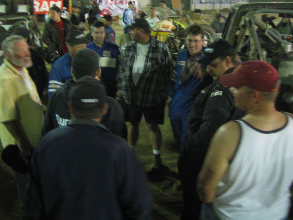 Crowd of men conversing at night beside an off-road vehicle in a dirt pit