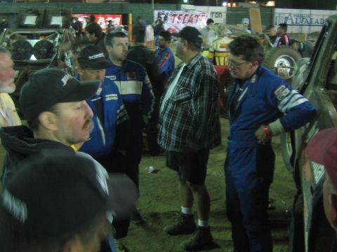 Men in blue racing suits and a plaid-shirted man talking among off-road cars at night