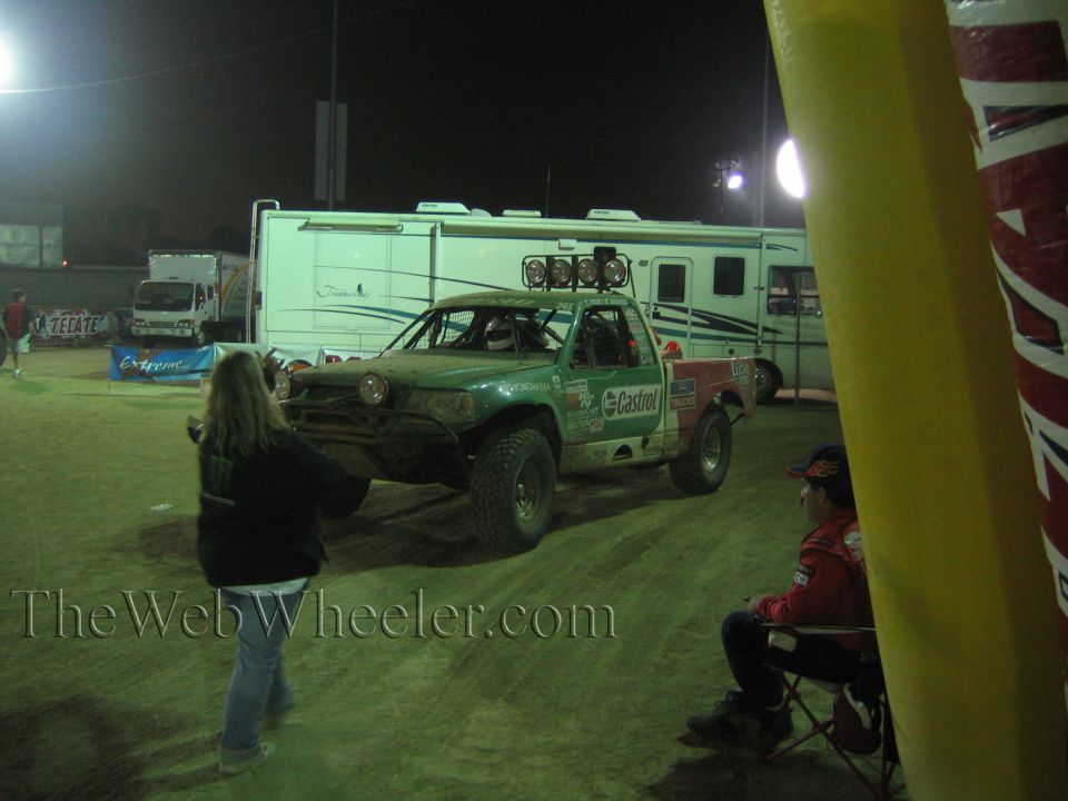 Green and white off-road race truck staged at night beside an RV motorhome