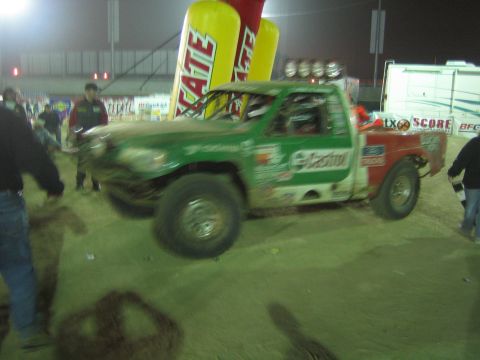 Green Castrol off-road race truck in motion on dirt at night near a Tecate banner