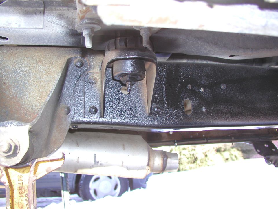 Underside view of a vehicle's frame with exhaust pipe visible. A jack stand is supporting the frame