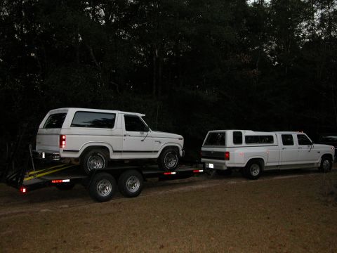 1986 Ford F-350 - A white Ford Bronco is loaded onto a trailer, while a white Ford F-350 crew cab dually with a camper shell is parked nearby.