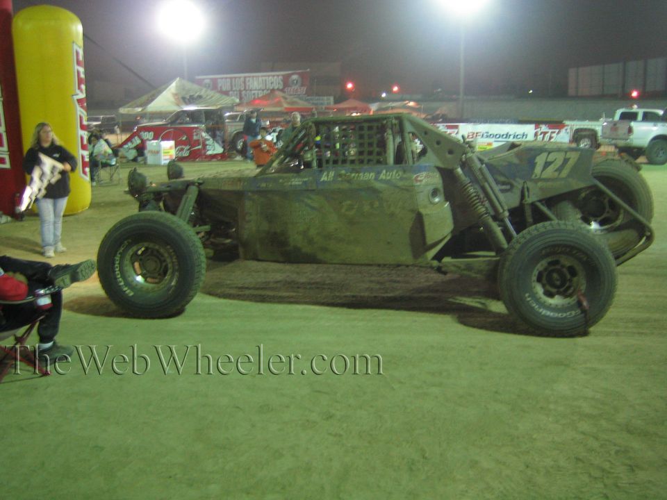 Mud-covered off-road buggy number 127 parked on dirt at night with people nearby