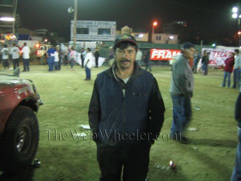 Man in dark jacket and cap standing in a dirt arena at night with race trucks