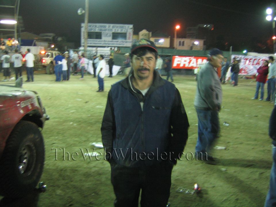 Man in dark jacket and cap standing in a dirt arena at night with race trucks
