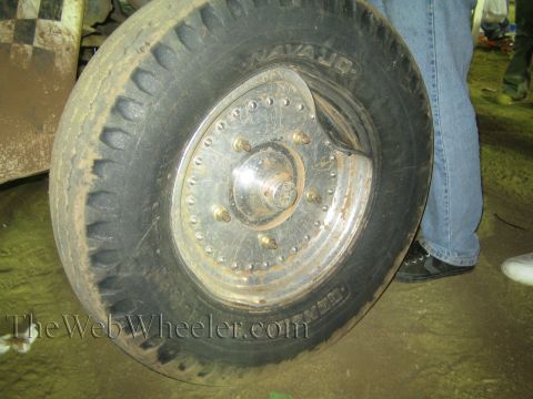 Close-up of an off-road tire and riveted metal wheel beside a person in jeans