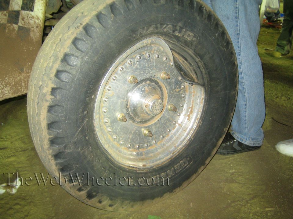 Close-up of an off-road tire and riveted metal wheel beside a person in jeans