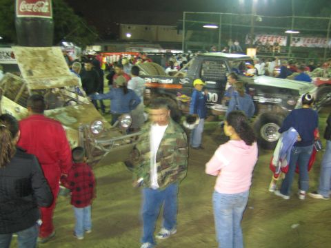 Busy crowd among off-road race vehicles in a dirt arena at night