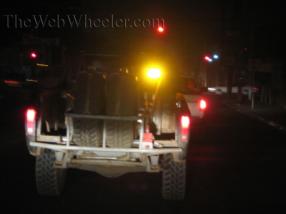 Rear of light-colored off-road truck on city street at night with amber and red lights