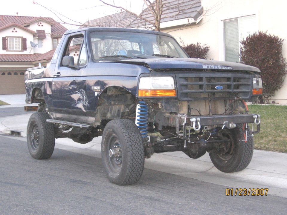 Lifted dark blue Ford Bronco with fenders and top removed, blue coil springs and tube bumper visible, parked on a street