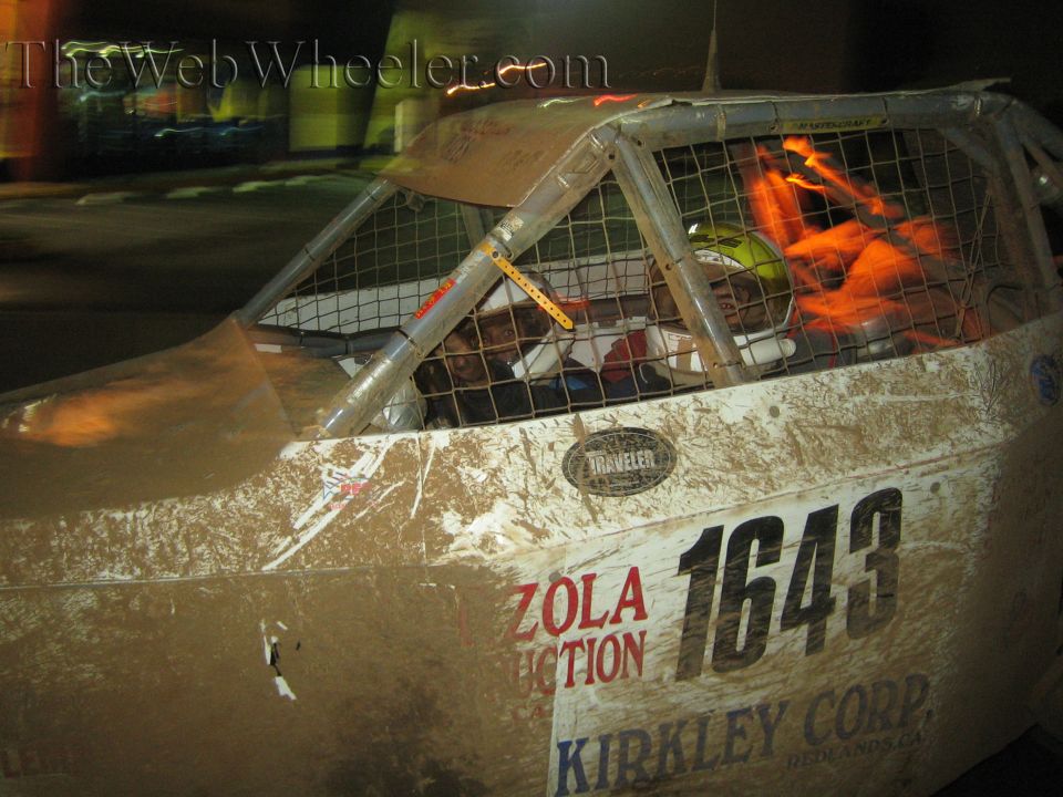 Side of muddy off-road race truck number 1643 with driver in orange suit in caged cab