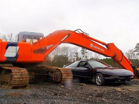 1993 Ford Probe - A black coupe is positioned next to a large orange excavator on a gravelly surface.