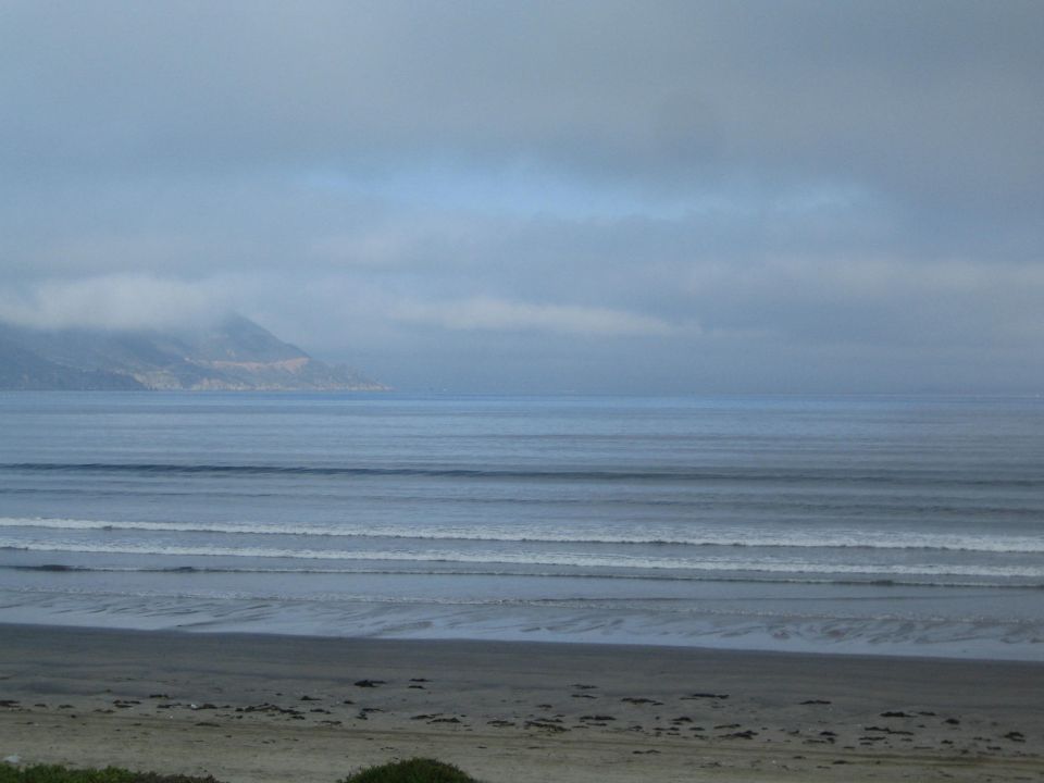 Overcast ocean view with rolling surf, sandy beach and fog-covered headland