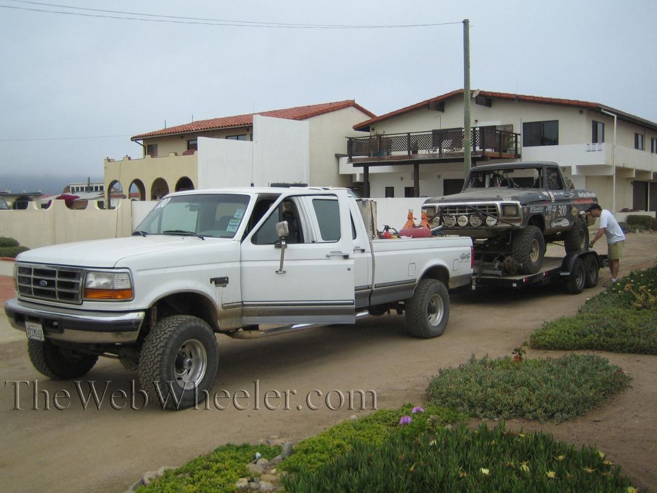 White Ford 4x4 pickup towing a trailer with an off-road truck by beach houses