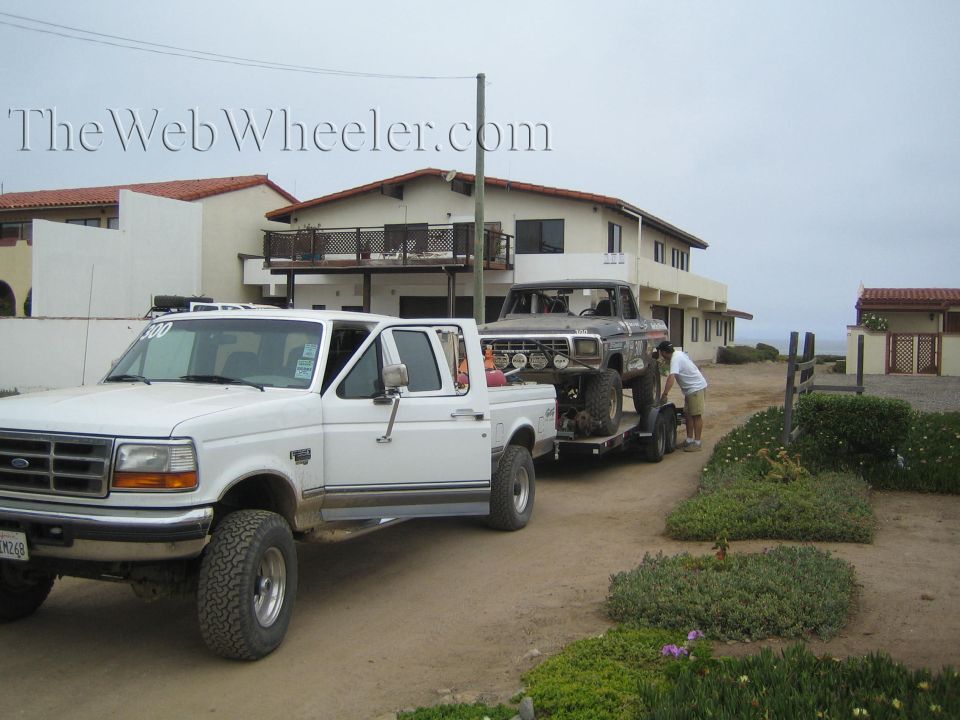 White Ford pickup towing an off-road truck on trailer in front of beach houses